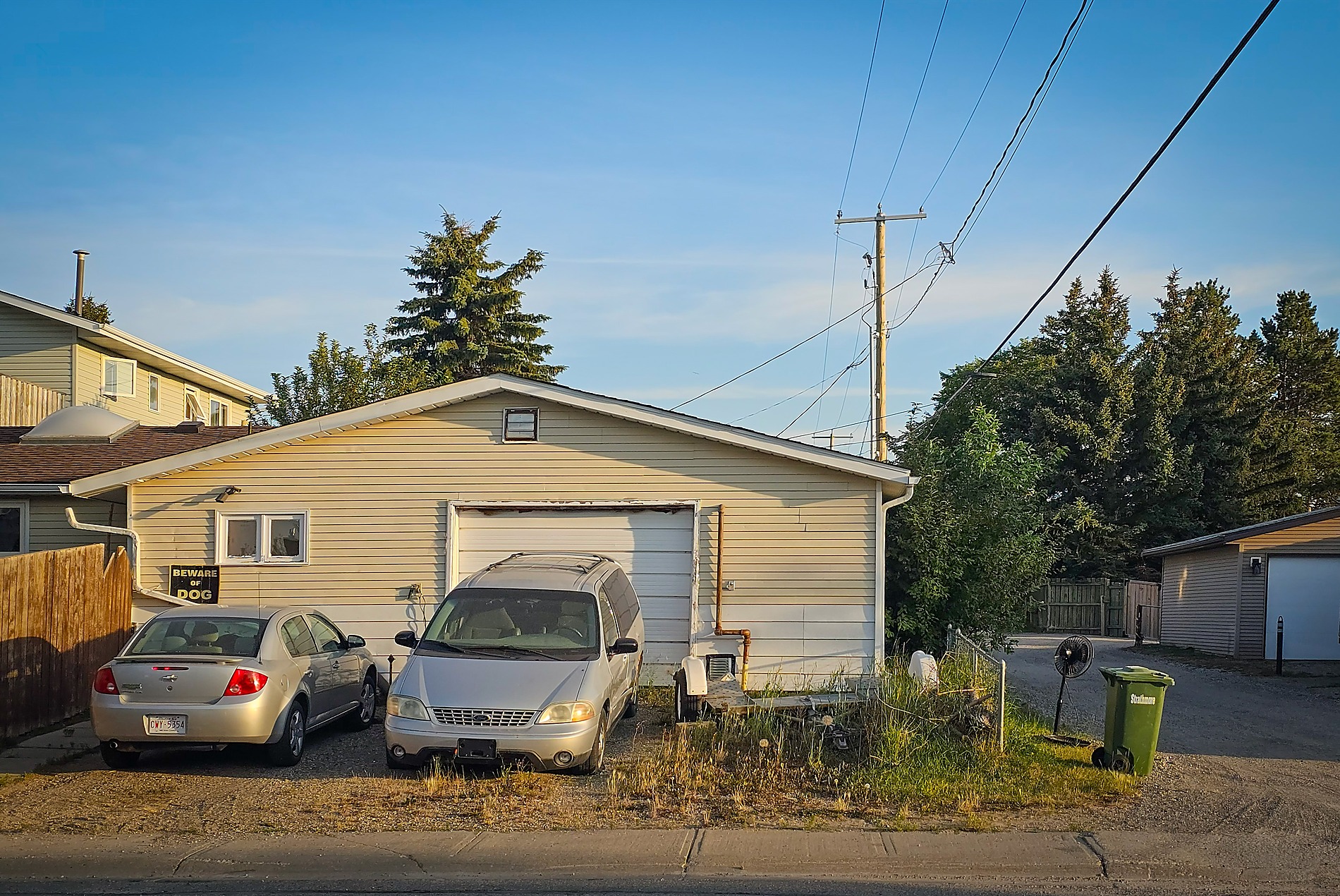 A cream-colored detached garage with a white garage door has a silver sedan and silver minivan parked on its weedy gravel driveway. A green garbage bin stands on the right next to a gravel alleyway, under a clear blue sky with utility wires and tall green evergreen trees.