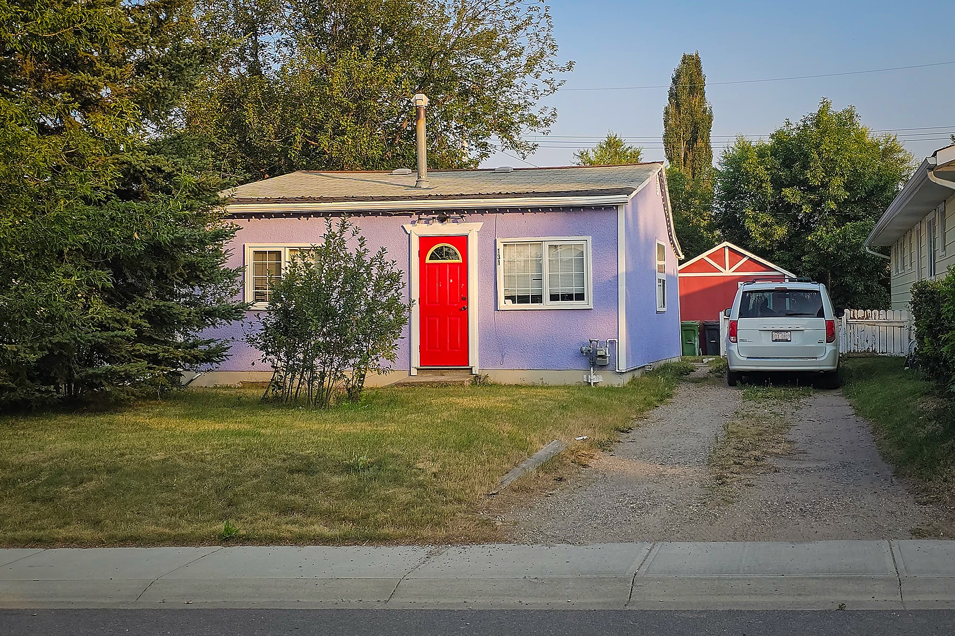 A small, one-story house with light purple stucco walls and a bright red front door is pictured from the street. A dirt and gravel driveway on the right holds a white minivan next to a small red shed.