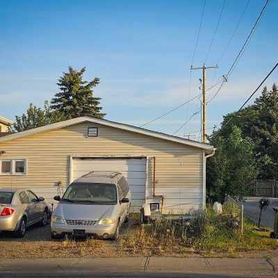 A cream-colored detached garage with a white garage door has a silver sedan and silver minivan parked on its weedy gravel driveway. A green garbage bin stands on the right next to a gravel alleyway, under a clear blue sky with utility wires and tall green evergreen trees.
