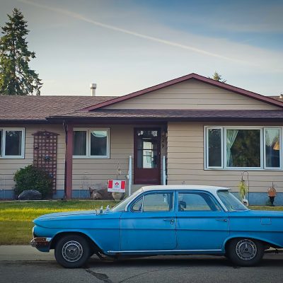 A vintage light-blue four-door sedan with a white top parked on an asphalt street in front of a tan bungalow with a brown-red roof, white windows, and a green grass lawn.