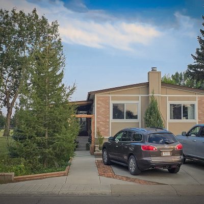 A single-story beige house with brick accents has a dark grey SUV and a silver pickup truck parked on its concrete driveway. A concrete path lined with evergreen trees leads toward the entrance under a blue sky with light clouds.