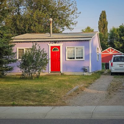 A small, one-story house with light purple stucco walls and a bright red front door is pictured from the street. A dirt and gravel driveway on the right holds a white minivan next to a small red shed.