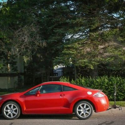 A side profile of a bright red two-door coupe parked on a paved street. Behind it, thick green trees and a hedge block the view of a residential house in the background.